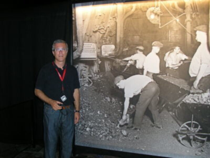 Persona posando junto a una imagen histórica de mineros trabajando en un entorno industrial.