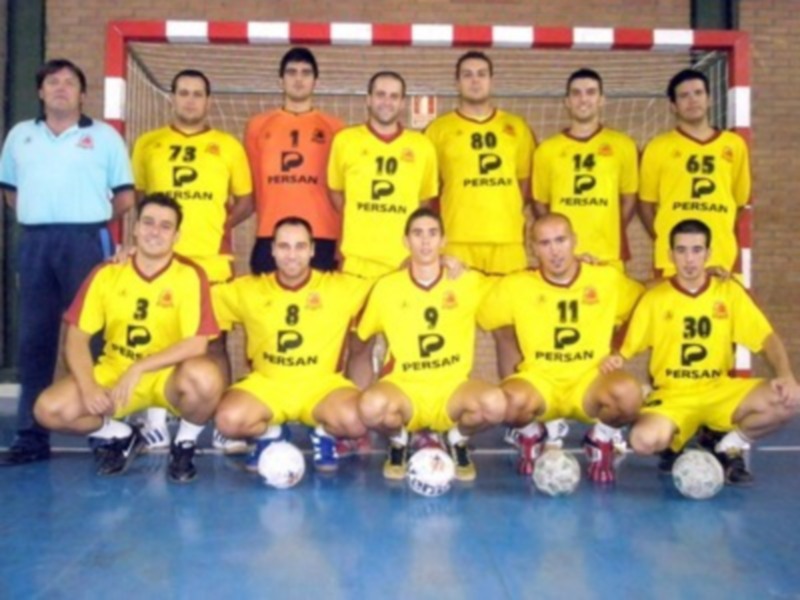 Equipo de balonmano con uniformes amarillos y pantalones cortos, posando con balones y entrenador detrás.