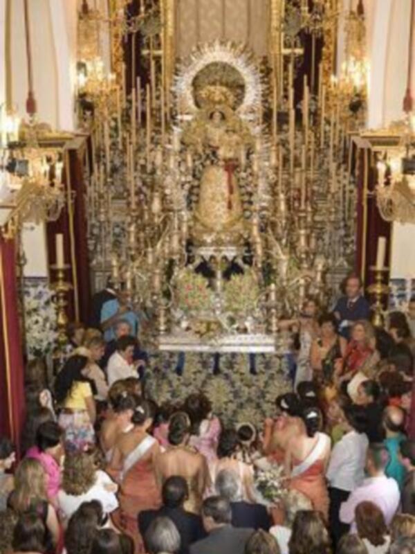 Una procesión religiosa con una imagen de la Virgen en el centro, rodeada de fieles en una iglesia decorada con luces y flores, celebrando una festividad religiosa.