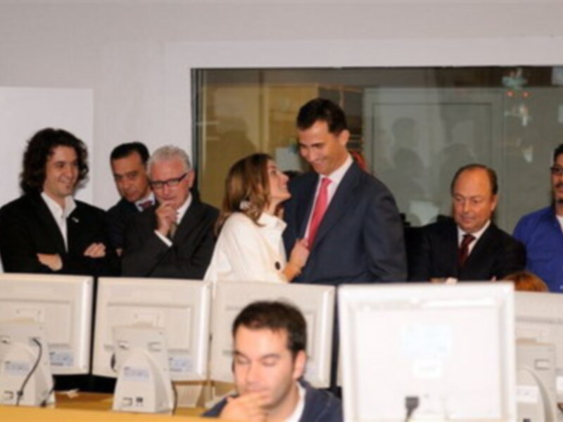 Una reunión formal con varios hombres y mujeres sentados en una sala de conferencias, enfocándose en una pantalla de computadora en el centro de la imagen.