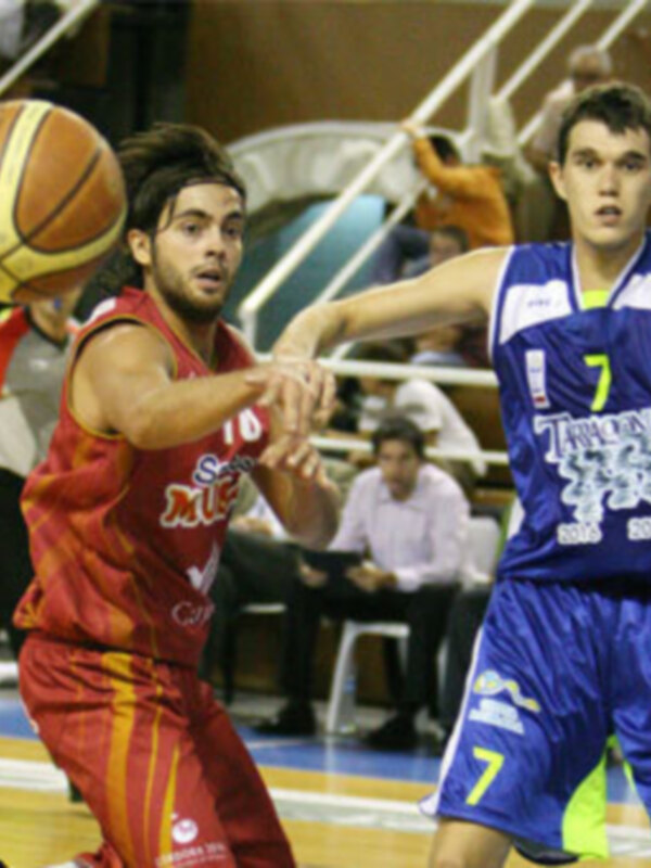 Jugadores en acción durante un partido de baloncesto, uno en rojo y otro en azul, en un entorno deportivo.