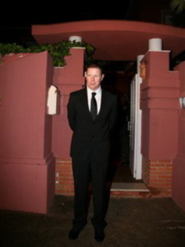 Un hombre en traje negro y corbata está de pie en una entrada de casa con una puerta abierta y una escalera al fondo. La iluminación es nocturna y el ambiente es formal.