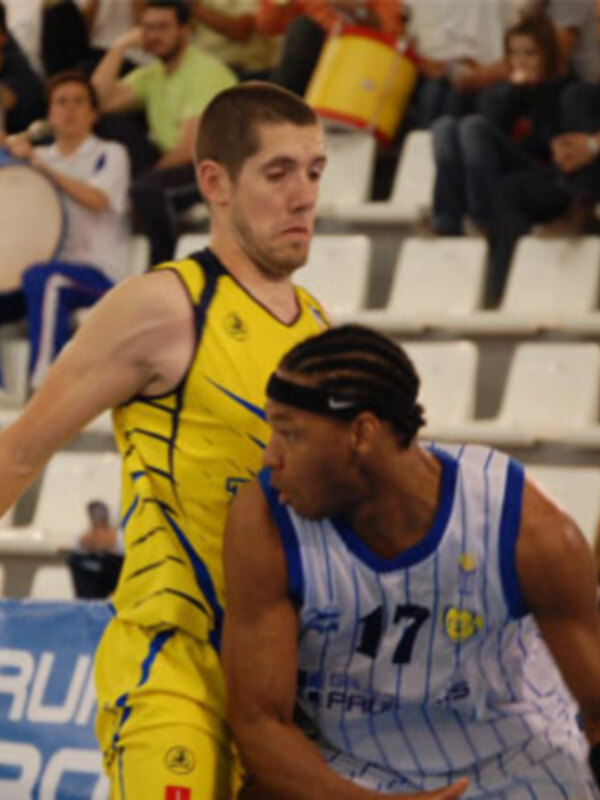 Jugadores de baloncesto en acción durante un partido, uno en amarillo y otro en azul, en un estadio con espectadores.