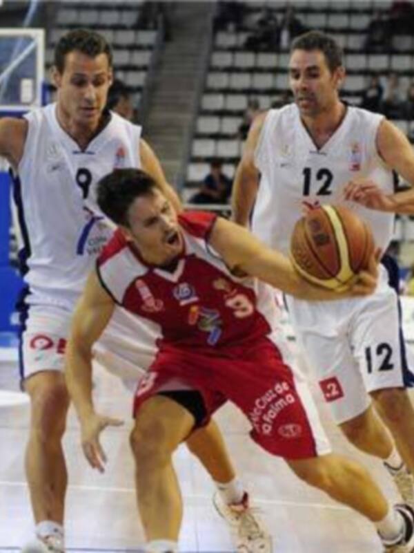 Jugador de baloncesto en acción durante un partido, con su equipo vestido de rojo y blanco, y el rival en blanco con el número 12. El fondo muestra una cancha de baloncesto con una grada vacía y un árbitro en el fondo.