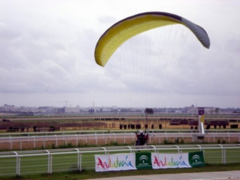 Parapente volando sobre una pista de carreras con banderas de Andalucía en primer plano.