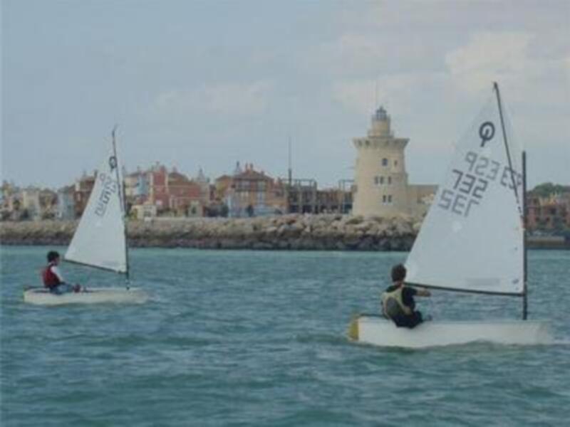 Dos regatistas en barcos a vela frente a un faro en el puerto.