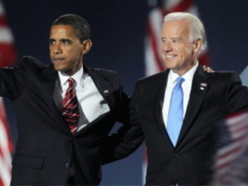 Obama y Biden en una reunión política, con banderas estadounidenses al fondo.