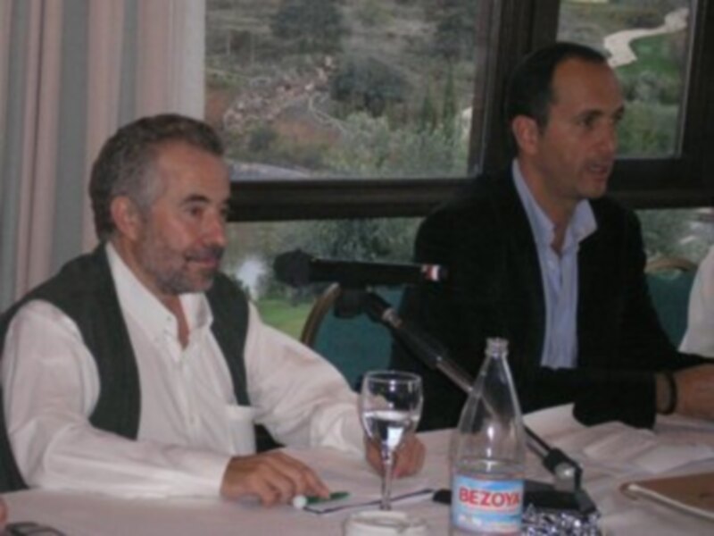 Dos hombres sentados en una mesa con microfonos, discutiendo en un evento público.