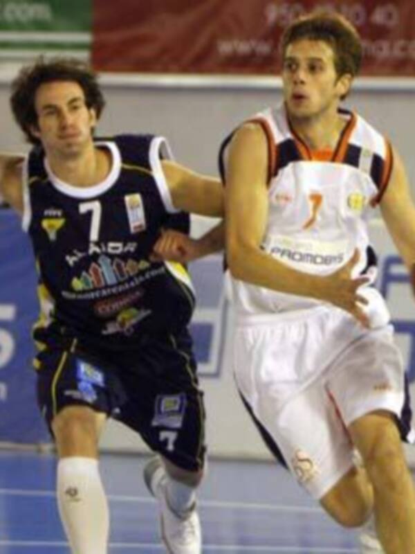 Jugadores de baloncesto en acción durante un partido, uno en azul y otro en blanco, corriendo por el balón en un piso de madera.