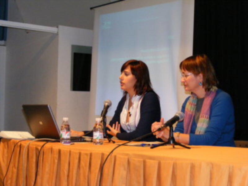 Conferencia con dos mujeres hablando en un panel, con una pantalla de proyección en el fondo y un ordenador portátil en la mesa.