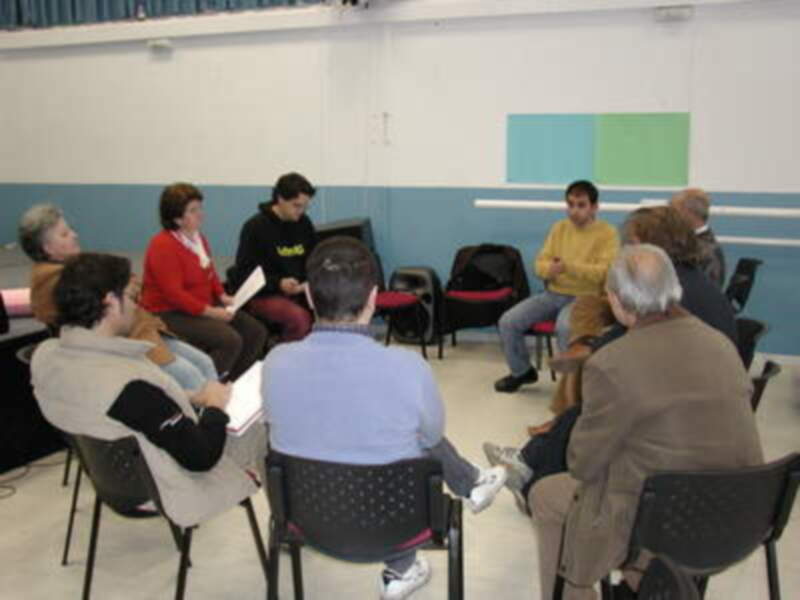 Una reunión de grupo en un aula con personas sentadas en sillas, discutiendo activamente. El ambiente es informal y colaborativo.