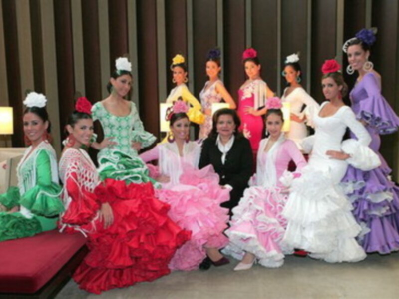 Una fila de mujeres vestidas con trajes de baile flamenco de colores vivos, incluyendo verde, rojo, rosa, blanco y púrpura, posando en un entorno elegante con cortinas marrones y una silla roja en el fondo.