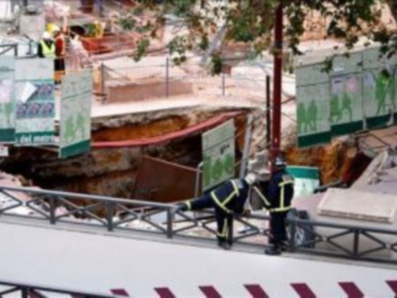 Trabajadores inspeccionan un pozo abierto en una calle urbana, con carteles informativos y una estructura de apoyo en el fondo.