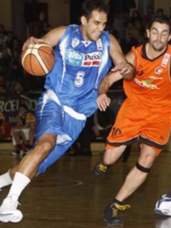 Jugadores de baloncesto en acción durante un partido, uno en azul y otro en naranja, en un entorno deportivo con público en el fondo.