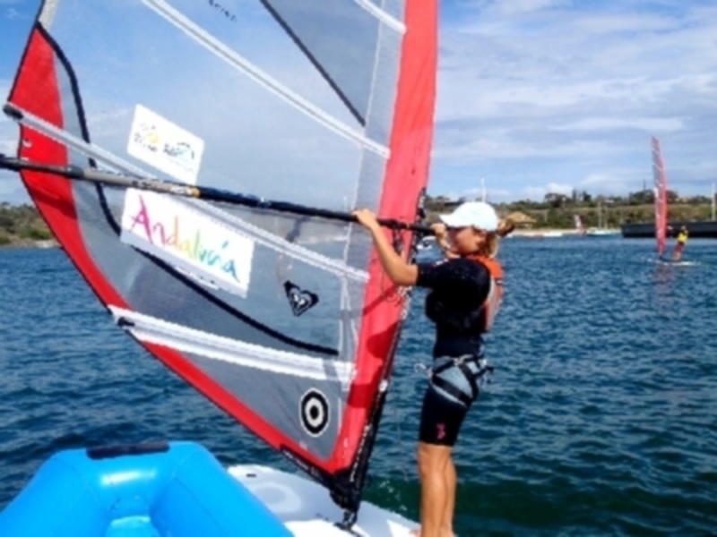 Una persona disfrutando de la navegación en un velero con una vela roja y gris, en un entorno de agua tranquila y cielo azul con nubes.
