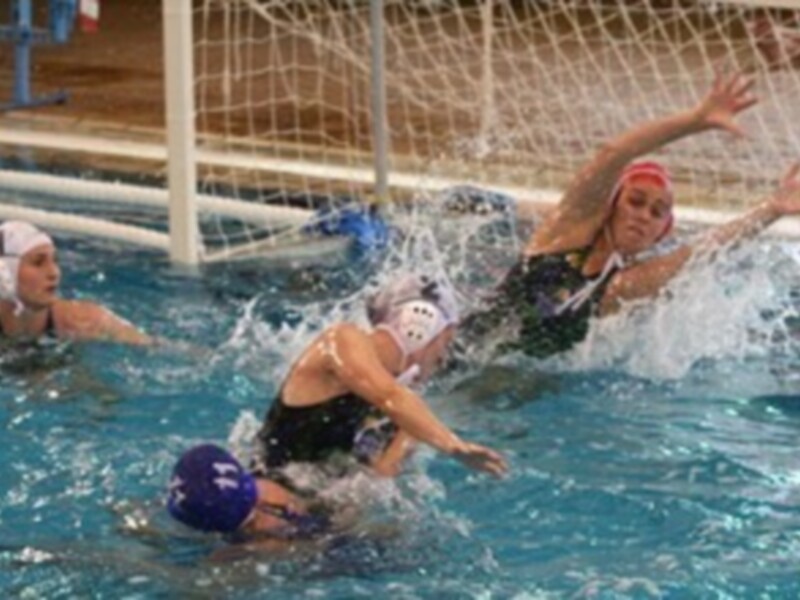 Jugadoras de waterpolo compiten en un partido en una piscina. Una jugadora lanza la pelota mientras otras se preparan para interceptarla.