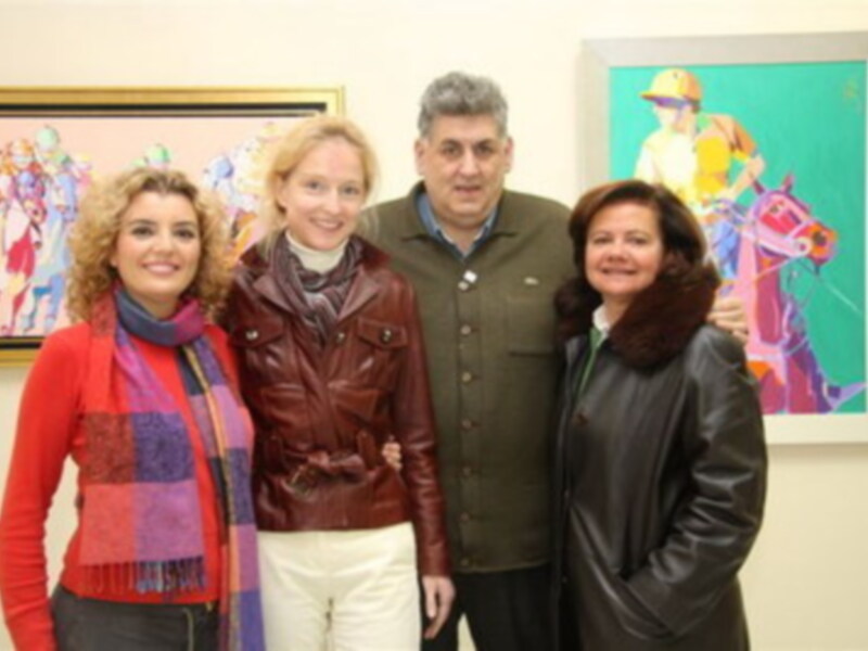 Una familia sonríe en una sala de exposiciones, con dos cuadros de arte en el fondo. La familia está compuesta por una mujer en el centro, una mujer a su izquierda y un hombre a su derecha. Los cuadros muestran personajes animados en colores vibrantes.