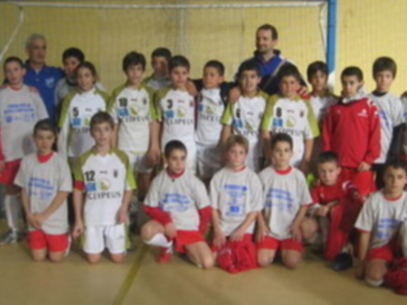 Un grupo de niños vestidos con camisetas blancas y rojas posan para una foto en un gimnasio. Algunos niños llevan medallas al cuello. La imagen muestra a los niños en una posición formal, posiblemente después de un entrenamiento o competencia.