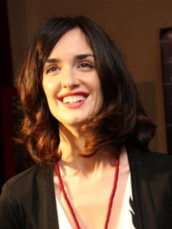Una mujer con cabello largo y rojo, sonriendo y mirando hacia la cámara. Se puede ver una parte de su rostro y parte de su vestimenta, que incluye una blusa blanca y una chaqueta negra. El fondo está oscuro, lo que destaca su rostro y su sonrisa.