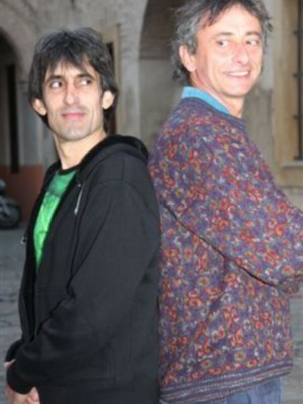 Dos hombres sonrientes posando juntos al aire libre, uno con chaqueta negra y otro con suéter estampado.