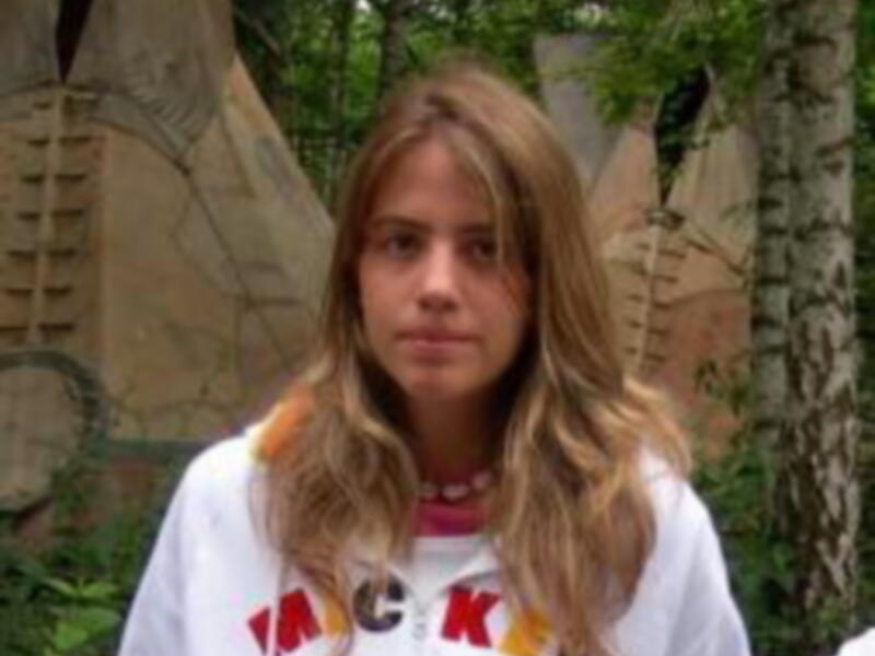 Una persona con cabello largo y rizado, llevando una camiseta con la palabra 'Racism' en el pecho, posando frente a un paisaje de rocas y vegetación.