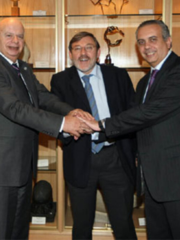 Tres hombres en trajes intercambian un apretón de manos en un entorno formal, posiblemente una reunión o ceremonia oficial.