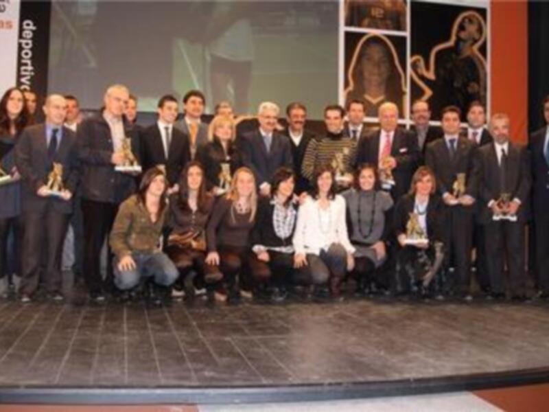 Ganadores de los premios de deporte en una ceremonia formal. Los ganadores posan con sus trofeos en un escenario con iluminación brillante y un fondo con imágenes deportivas.