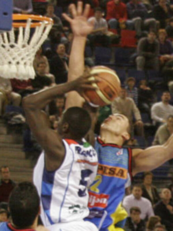 Jugadores de baloncesto en acción durante un partido, uno intenta marcar mientras el otro bloquea el balón.