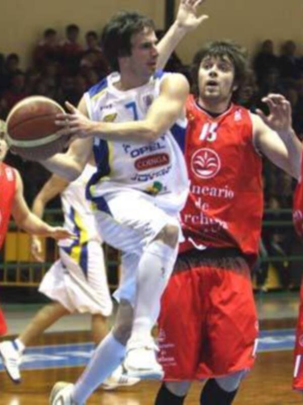 Jugadores de baloncesto en acción durante un partido, uno en blanco y otro en rojo luchando por el balón.