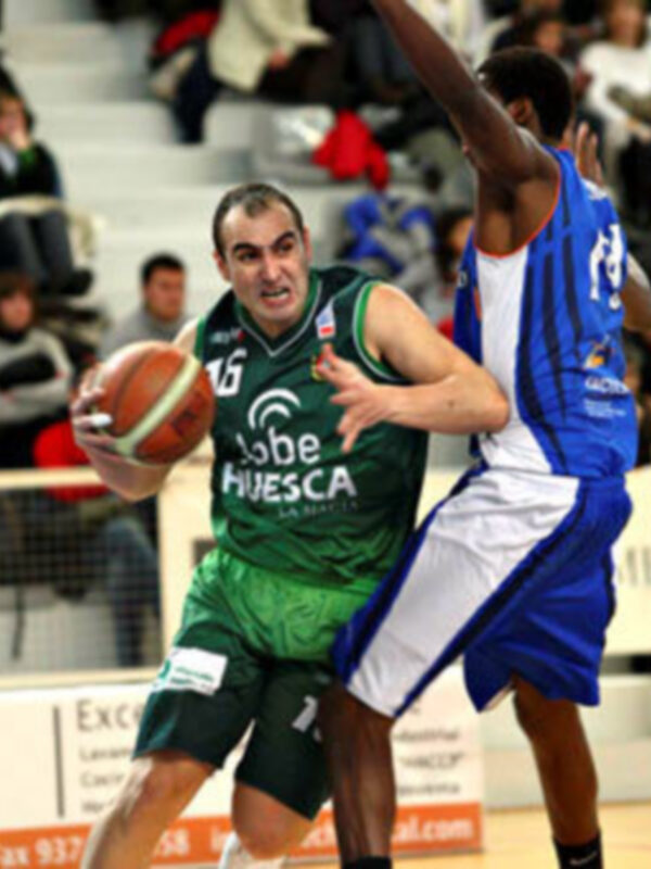 Jugadores de baloncesto en acción durante un partido, uno en verde y otro en azul, en un entorno deportivo con público en el fondo.
