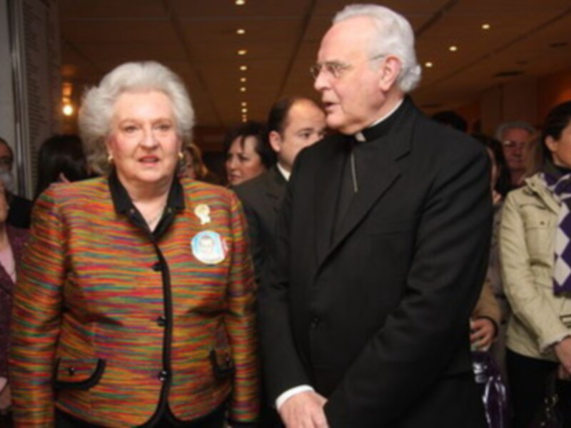 Una imagen de una mujer mayor con un abrigo colorido y una medalla de la paz, caminando junto a un sacerdote en una reunión formal.