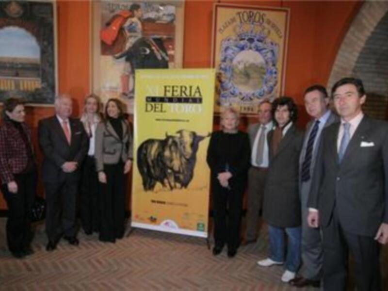 Una imagen de una feria de toros en España, con personas en trajes formales posando frente a un cartel de la feria. El ambiente es formal y festivo, con decoración típica de ferias taurinas.