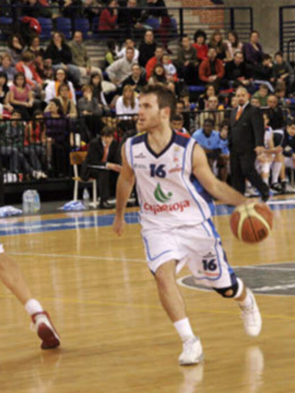 Jugador de baloncesto en acción durante un partido, driblando el balón con determinación en un entorno deportivo con público en el fondo.