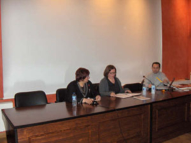 Reunión de trabajo en sala de conferencias con tres personas sentadas en una mesa con documentos y microfonos.
