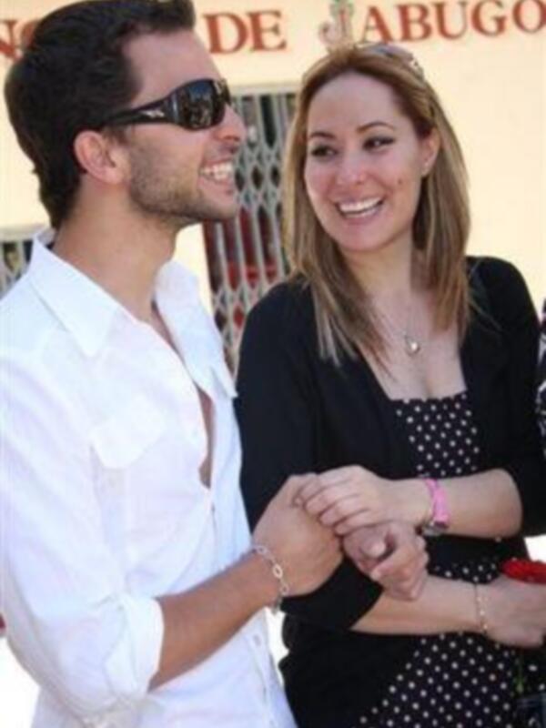 Una pareja sonriendo en una celebración al aire libre. La mujer lleva un vestido con lunares y una pulsera, mientras que el hombre está sin camisa y con gafas de sol. El fondo muestra un cartel con texto en español.