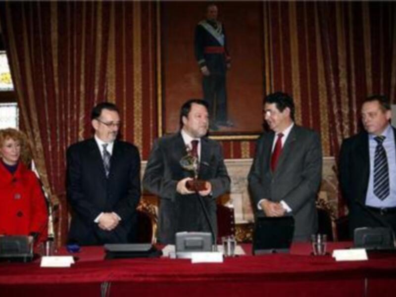 Una ceremonia formal donde un hombre sostiene un premio mientras otros lo felicitan. El escenario es elegante con cortinas y una estatua en el fondo. Los asistentes están vestidos formalmente, lo que sugiere una celebración o conmemoración importante.