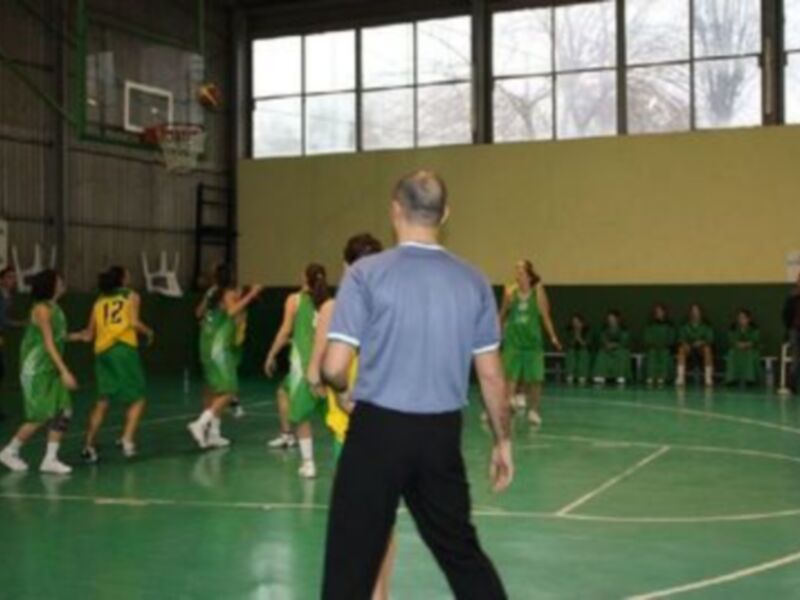 Juego de baloncesto en un gimnasio con jugadores en uniformes verdes y un árbitro en el centro.