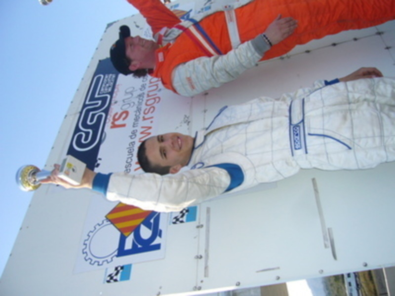 Un piloto de carreras posa en el podio con una medalla en la mano, sosteniendo una bandera nacional y un trofeo. El fondo muestra la bandera nacional y el logotipo de la competición.