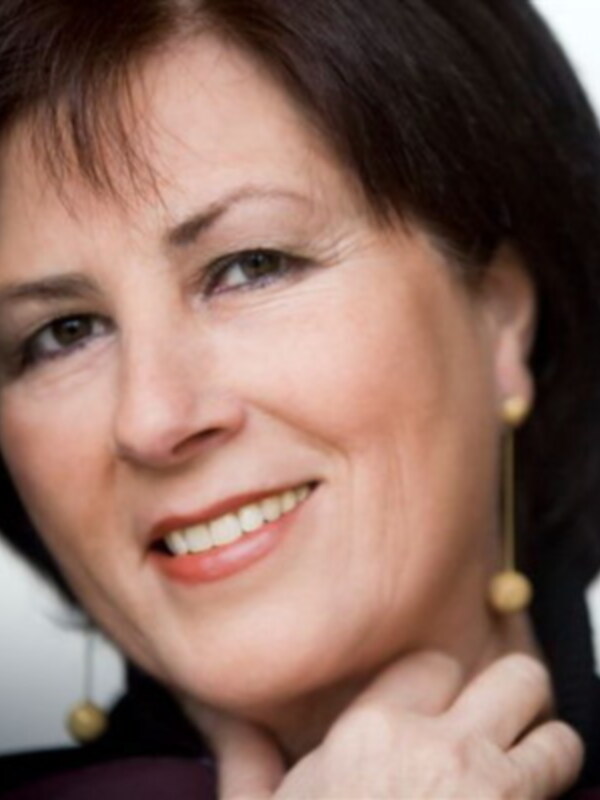 Una mujer con cabello oscuro cortado hasta la nuca, sonriendo con una expresión amable. Está usando un vestido oscuro y pendientes dorados. El fondo es blanco y suave, enfocándose en su rostro y expresión.