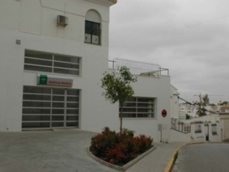 Edificio blanco con garaje y flores en el jardín frontal, ubicado en una zona urbana con edificios adyacentes.