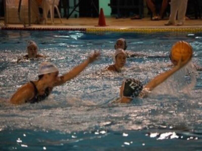 Un grupo de nadadores en acción en una piscina de agua salada, con una persona en el centro del agua usando un palo de waterpolo y otros nadadores alrededor.