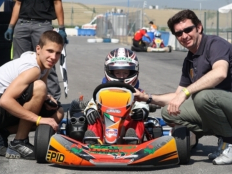 Un niño en una karting roja y naranja, con un piloto de carreras en el fondo, y un hombre en una camiseta negra y pantalones cortos, posando junto a la karting.