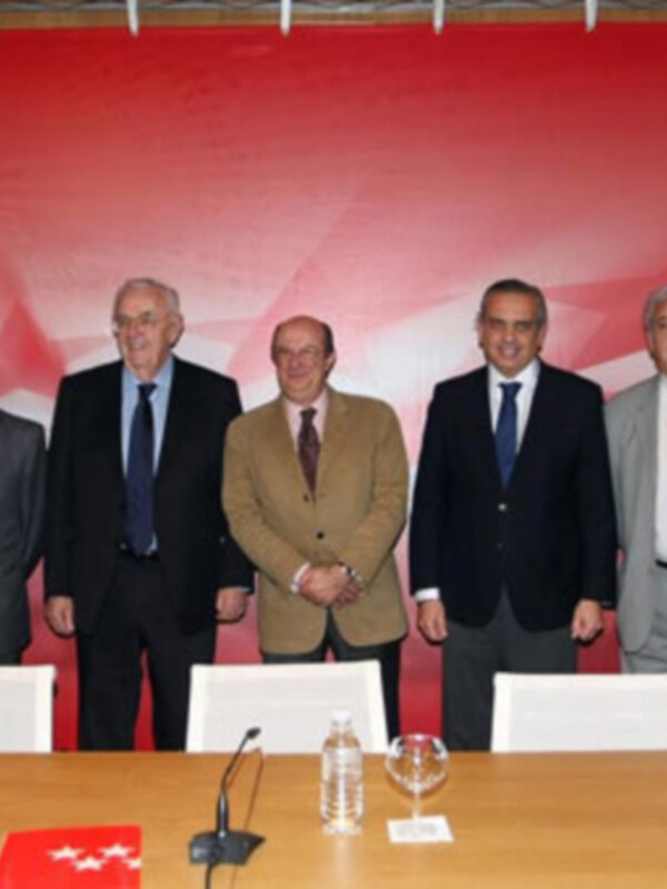 Un grupo de hombres en trajes posando para una foto en una reunión formal. El fondo es rojo con una bandera visible. Hay un microfono, vasos de agua y una bandera en el escritorio. El ambiente sugiere una reunión de negocios o política.