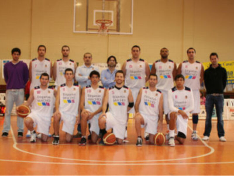 Equipo de baloncesto posando en el aula de entrenamiento, con uniformes blancos y rojos, frente a un fondo de pared amarilla y una cancha de baloncesto. El equipo parece estar listo para una competición o un partido.