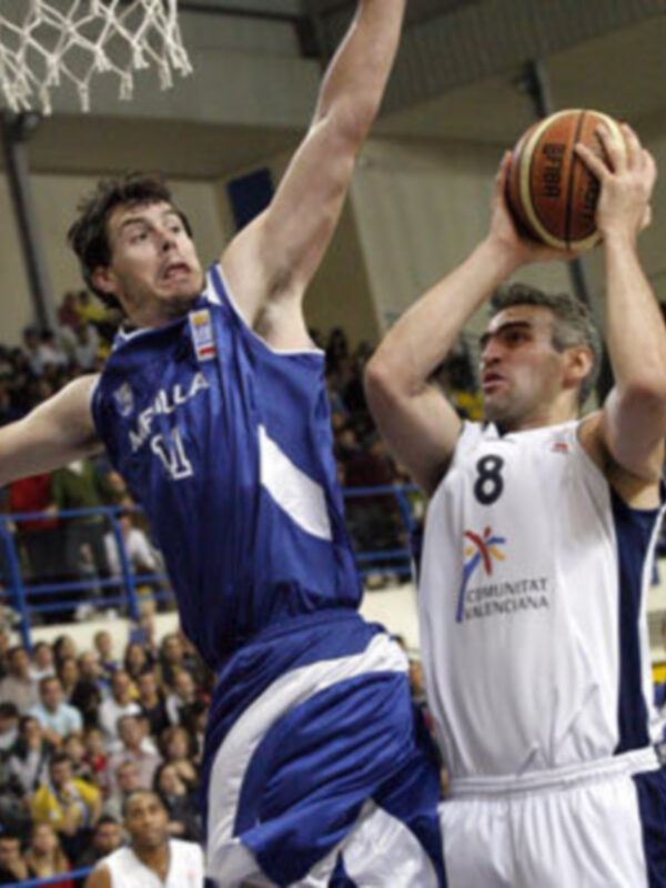 Jugadores de baloncesto en acción durante un partido, uno intentando hacer un tiro y el otro defendiendo.