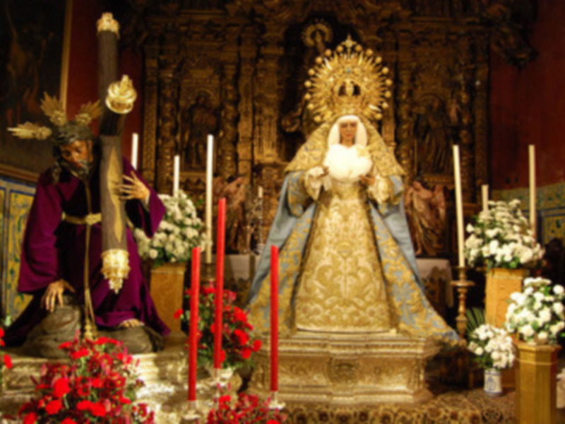 Imagen de una iglesia con dos estatuas religiosas, una vestida de negro y la otra de dorado, rodeadas por velas rojas y flores.