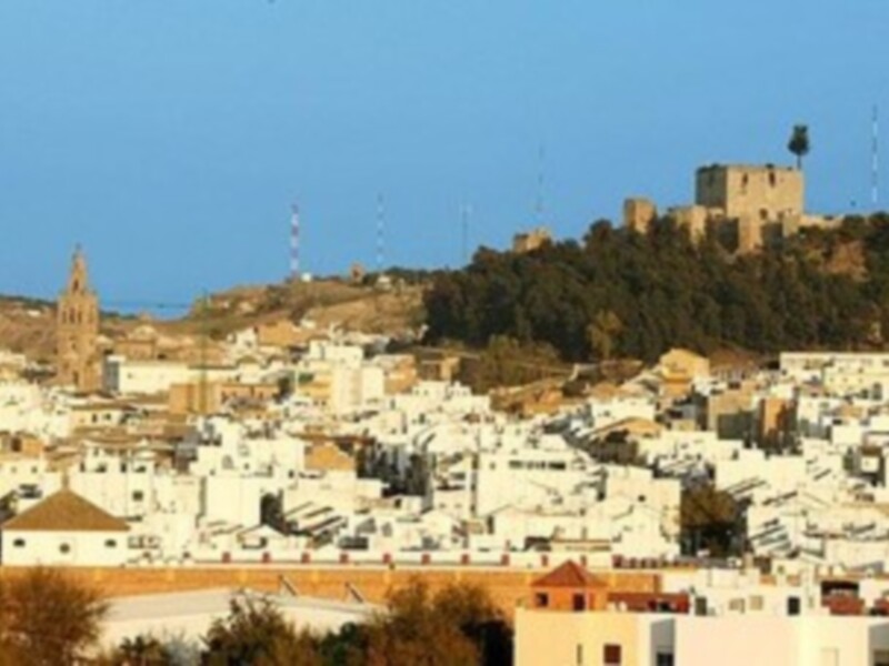 Vista panorámica de una ciudad con edificios blancos y una torre de iglesia en el horizonte, bajo un cielo azul claro.