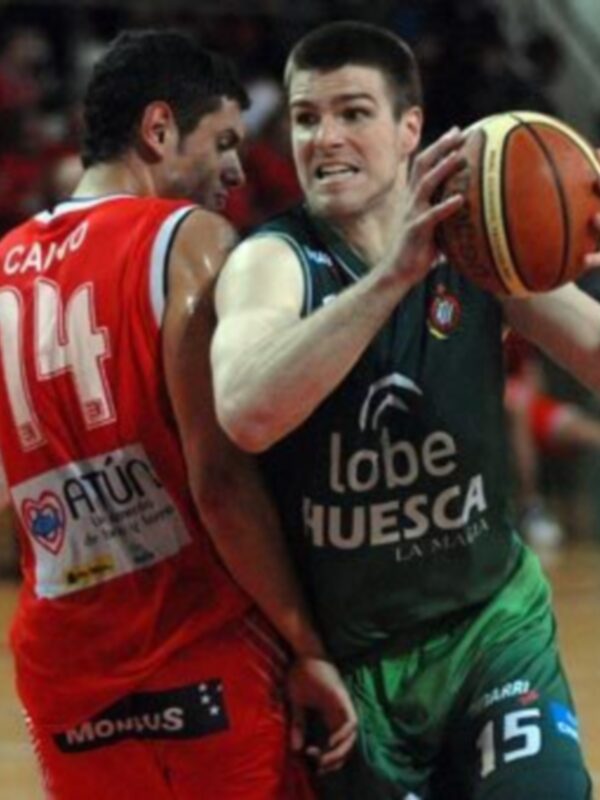 Jugador del equipo Huesca en acción durante un partido de baloncesto, controlando el balón bajo la defensa del rival en rojo.
