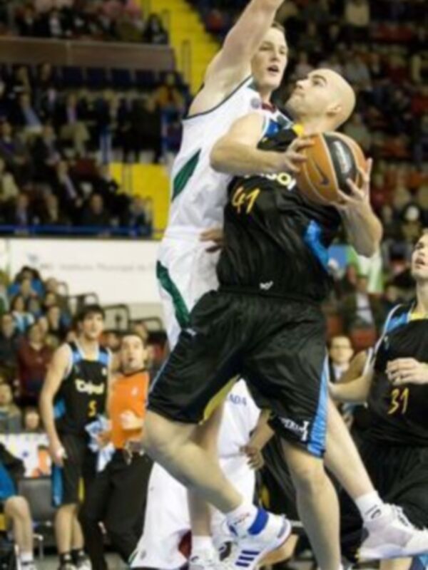 Un jugador de baloncesto en acción durante un partido, con el número 44 en el uniforme negro y blanco, intentando anotar mientras es defendido por un jugador del equipo contrario.