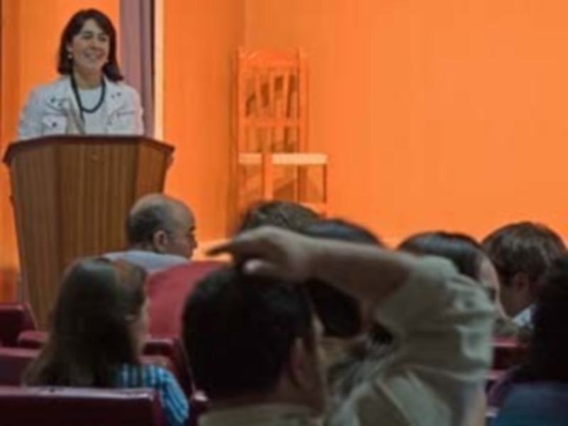 Lectura en escenario con público atento, fondo naranja y silla vacía.